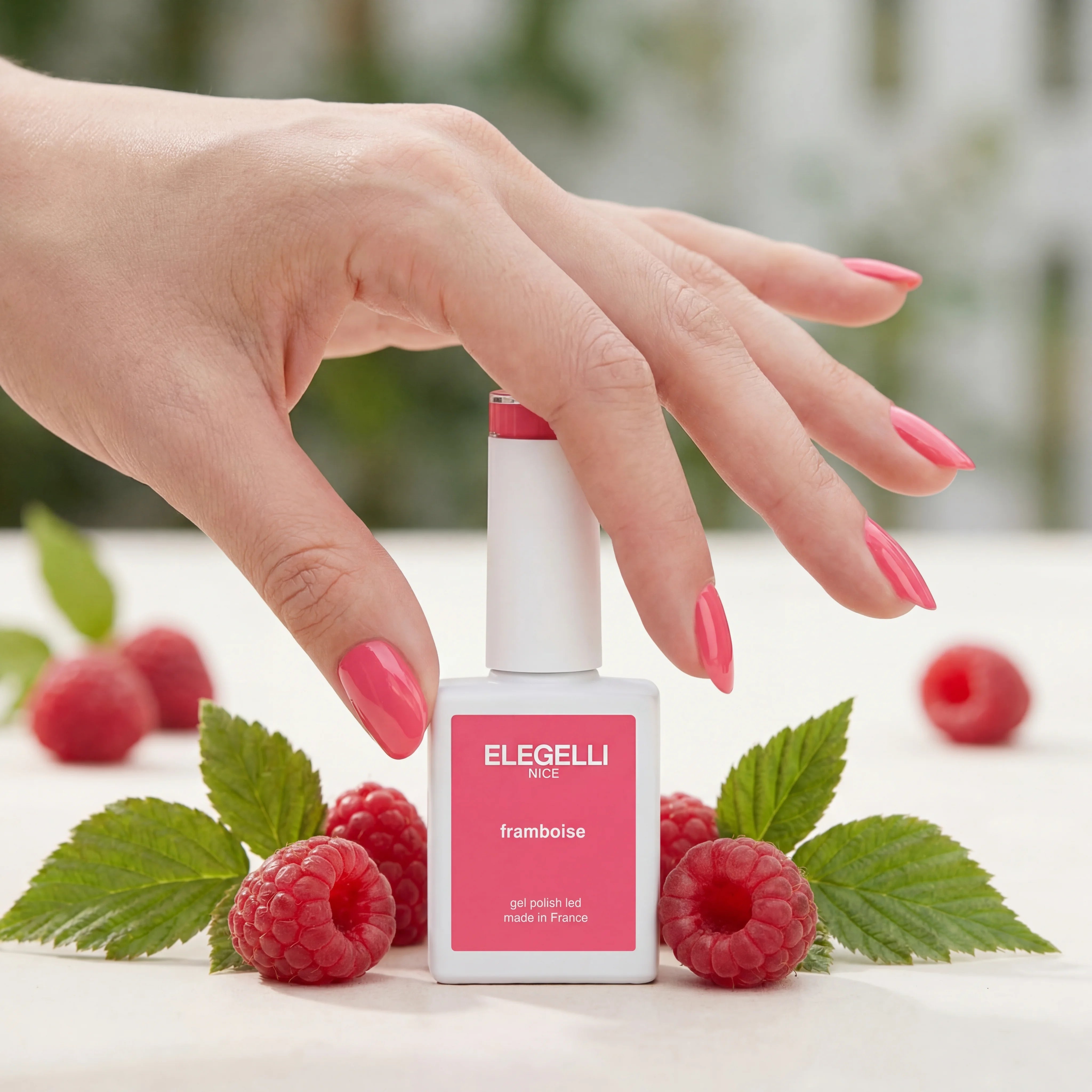 Hand with pink nail polish holding a bottle of Elegelli nail polish surrounded by raspberries and leaves.